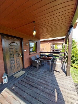 Relax on the covered deck with a wooden door, table, and chairs, surrounded by a railing with a view of the building.
