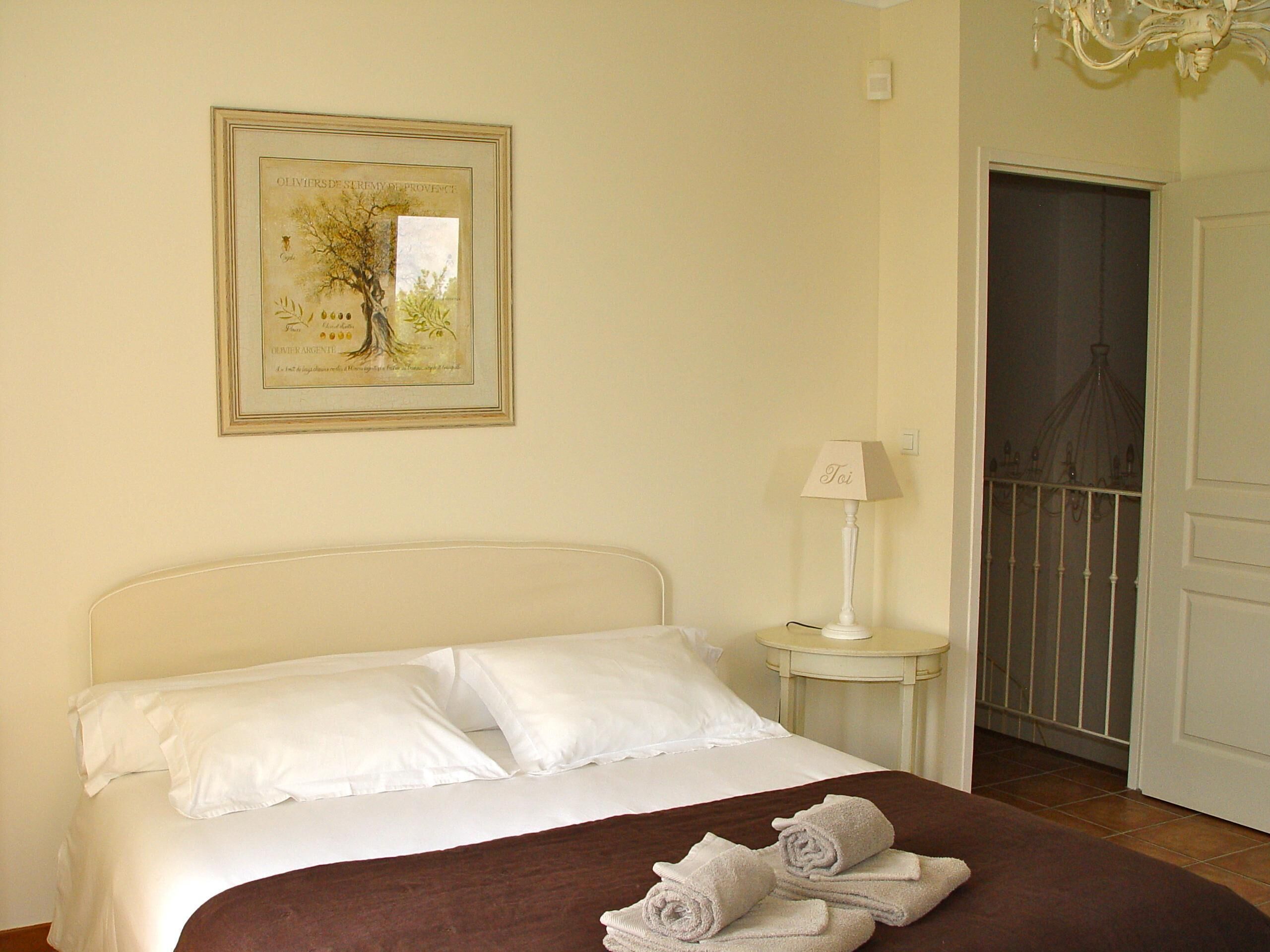 A hotel room with a bed, a nightstand, a lamp, a framed botanical illustration, and a door.