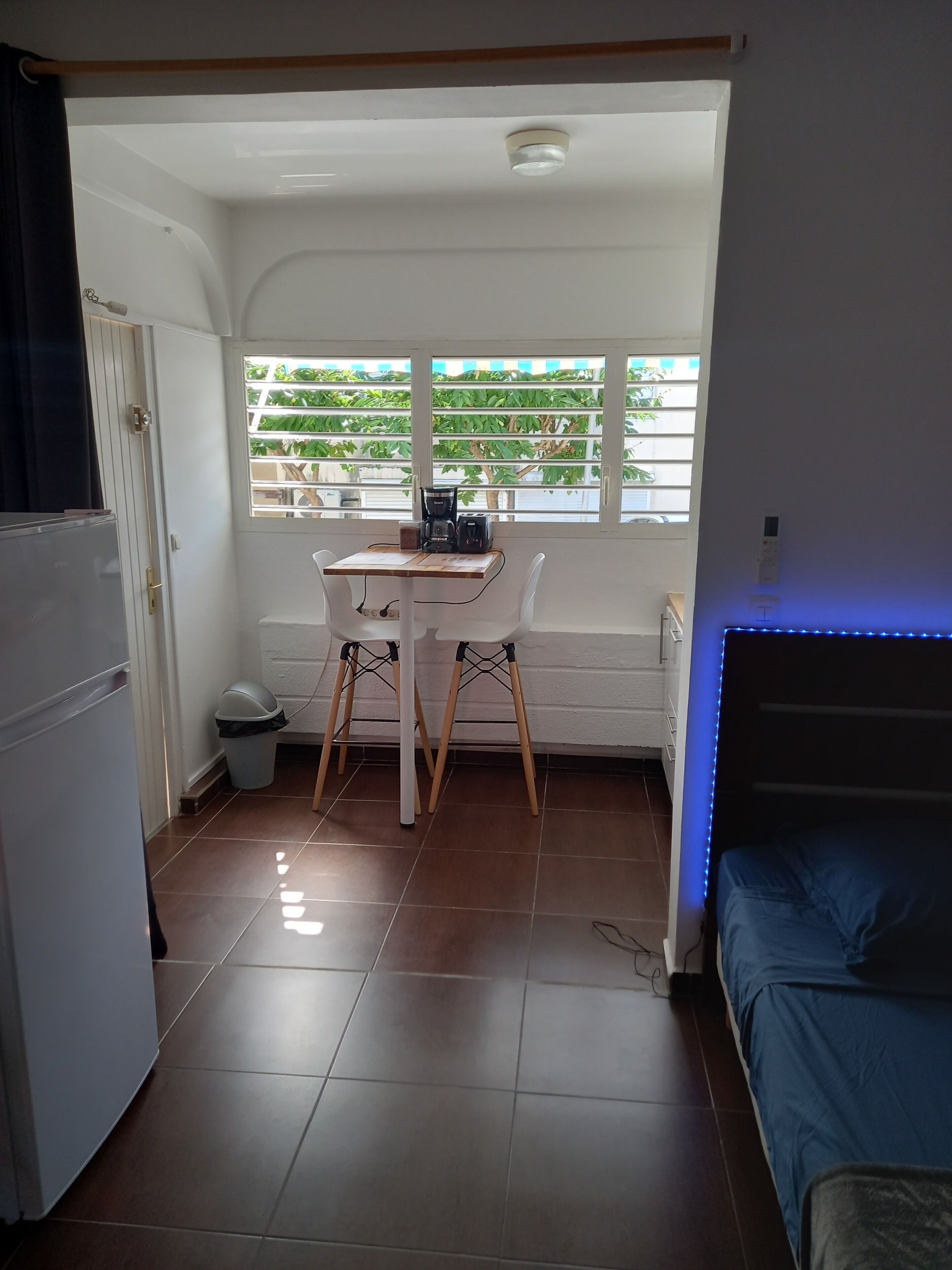 Una habitación pequeña con una mesita, dos sillas, una cama y un ventanal con persianas.