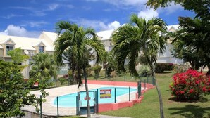 A pool area with a fenced deck, surrounded by palm trees and a building with a white roof. - Studio Oncle Sam: pool and beach access. Ideal for vacations or weekends for two (Le Gosier)