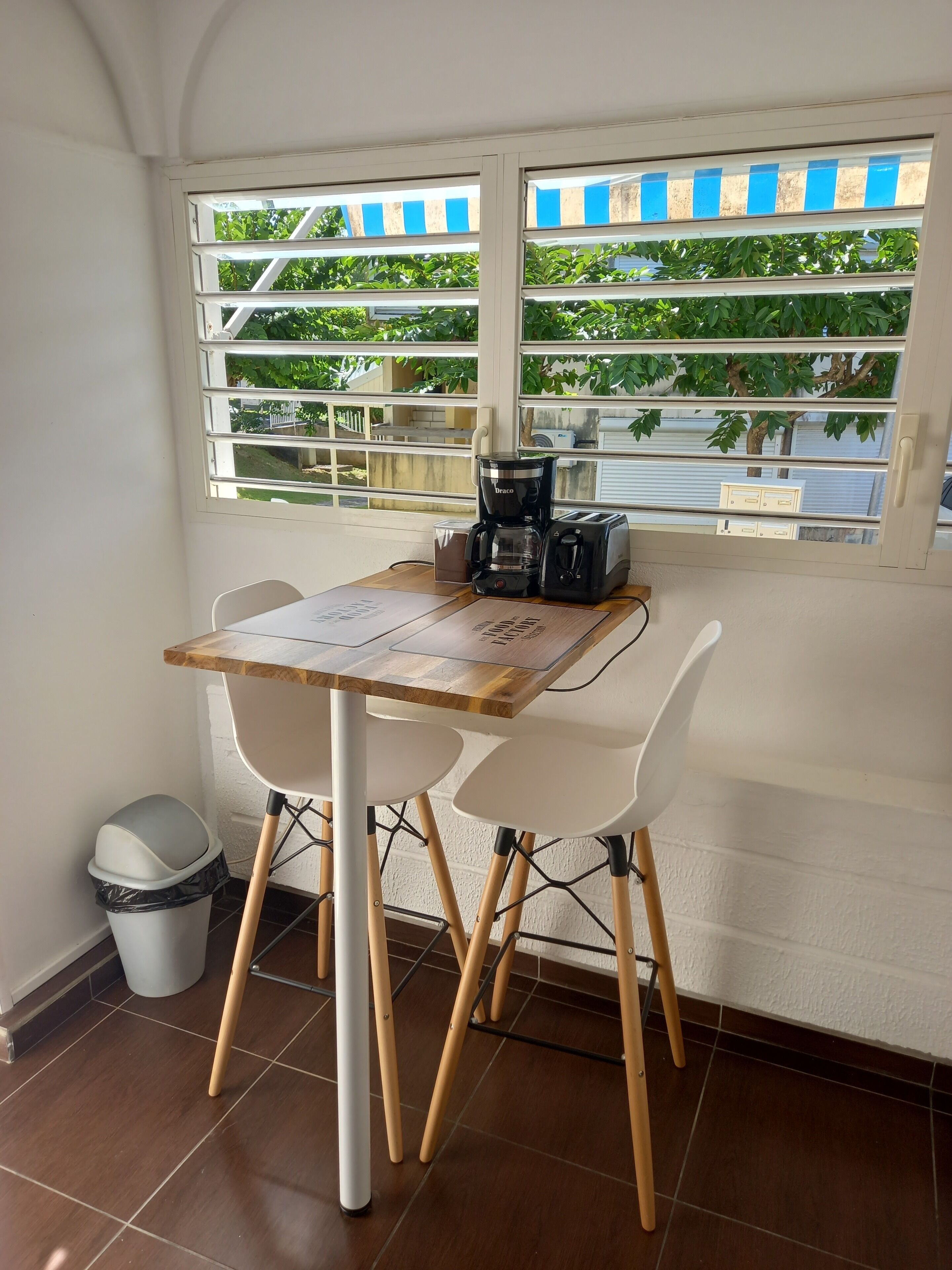 Un pequeño comedor con una mesa y sillas, una cafetera y una ventana con persianas.