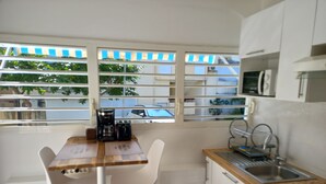 A compact kitchen with a wooden table, white chairs, a built-in microwave, and a window with a view of the street. - Studio Oncle Sam: pool and beach access. Ideal for vacations or weekends for two (Le Gosier)