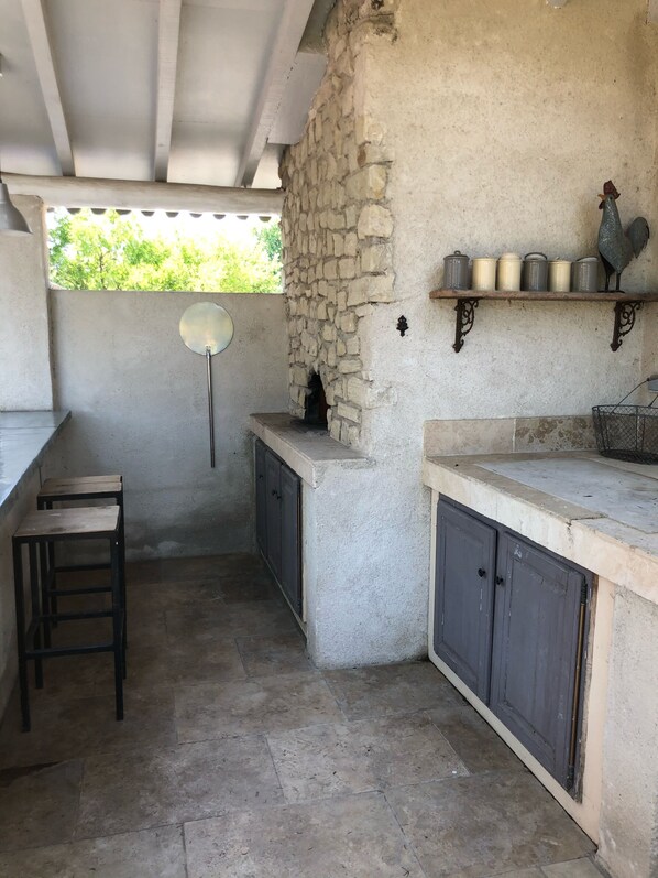 A rustic outdoor kitchen with a stone wall, wooden stools, and a shelf with kitchenware.