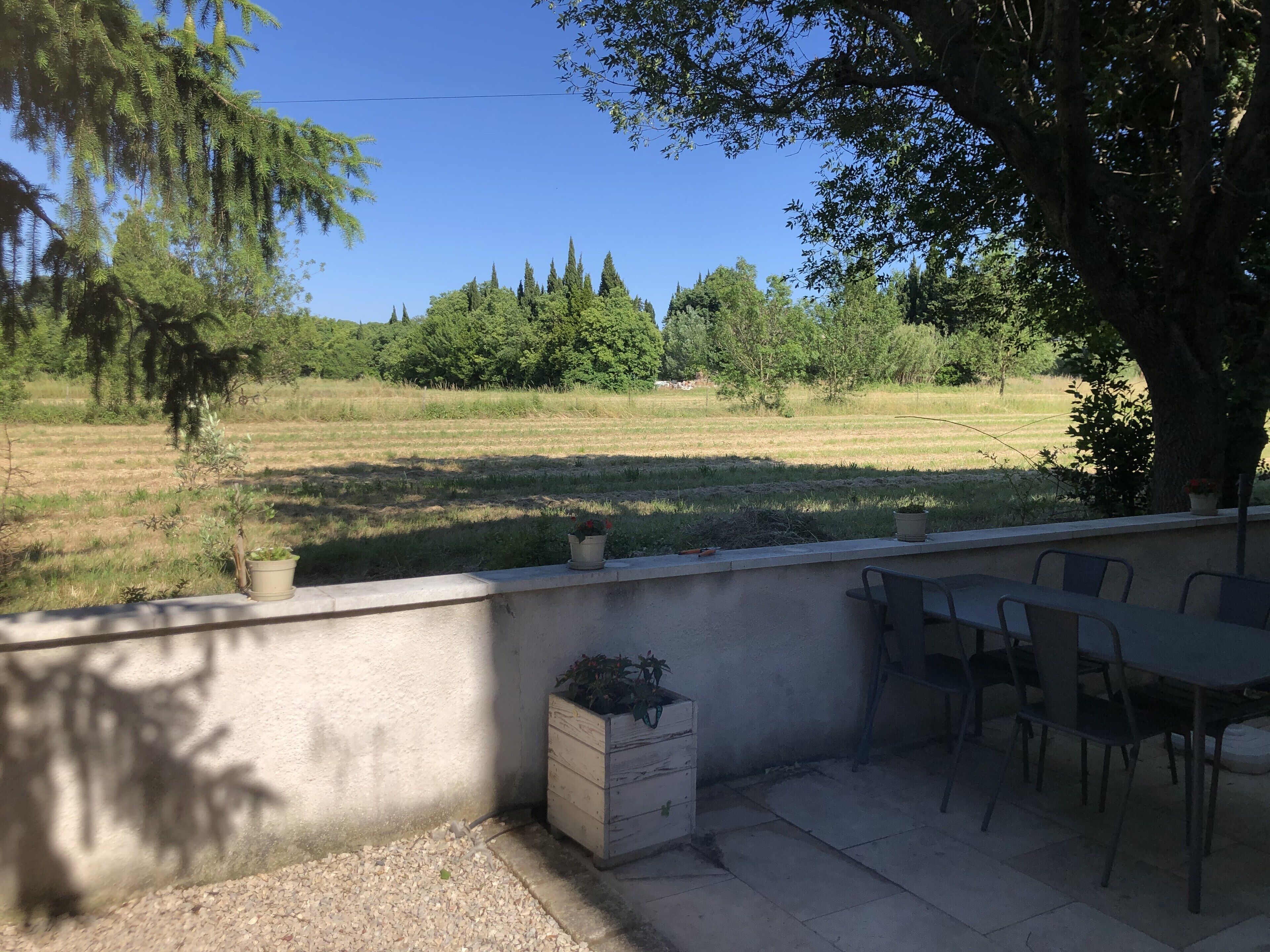 An outdoor patio area with a concrete wall, a table, and chairs. There are potted plants on the wall and a small wooden chest. The background features a grassy field and trees.