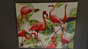 Une chambre à coucher avec un grand tableau représentant des flamants roses sur le mur, une tête de lit en bois et un lit recouvert d’une couette blanche.