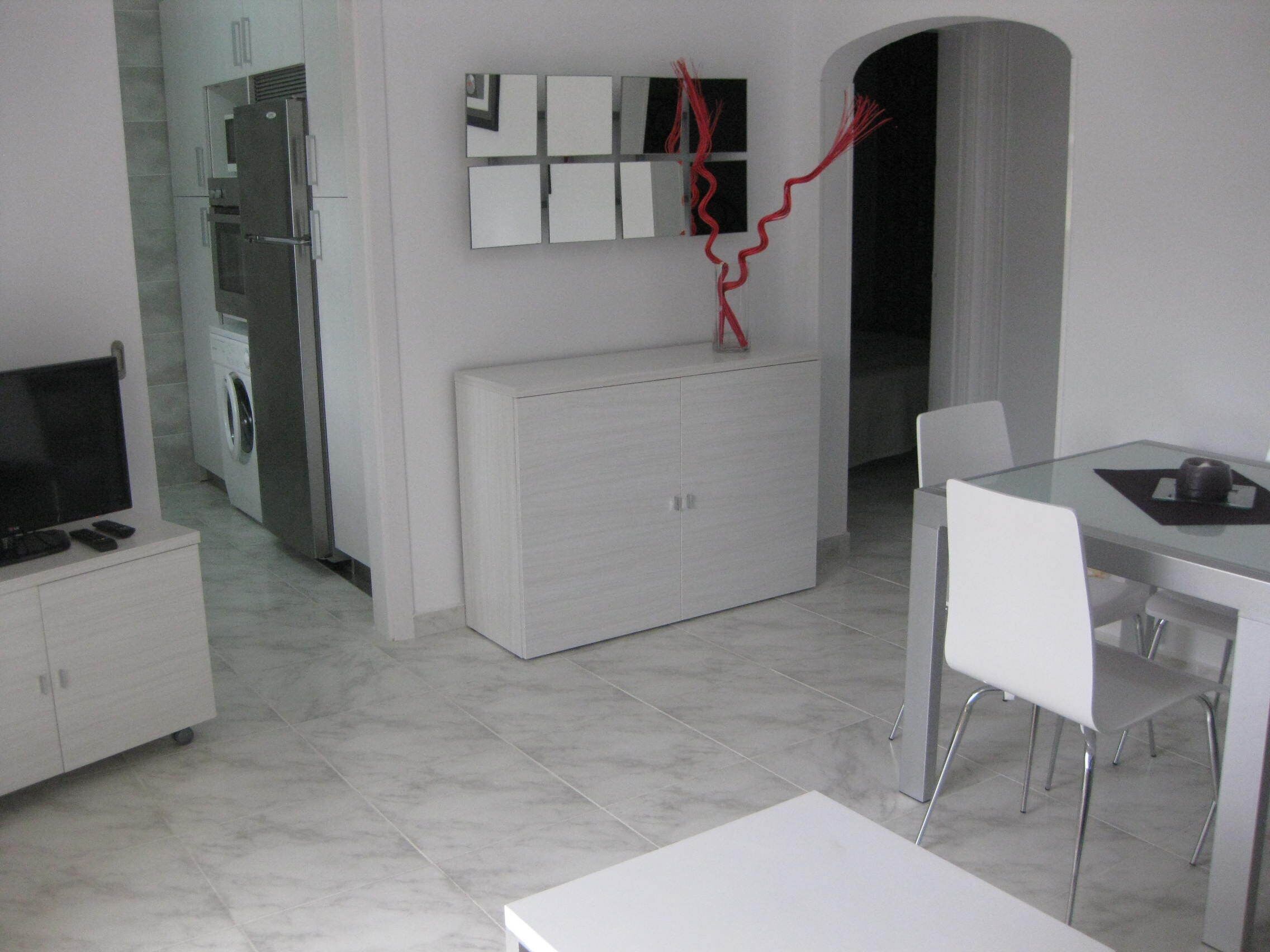 Una cocina moderna con horno empotrado, microondas y refrigerador. Hay un comedor con mesa de vidrio y sillas blancas.