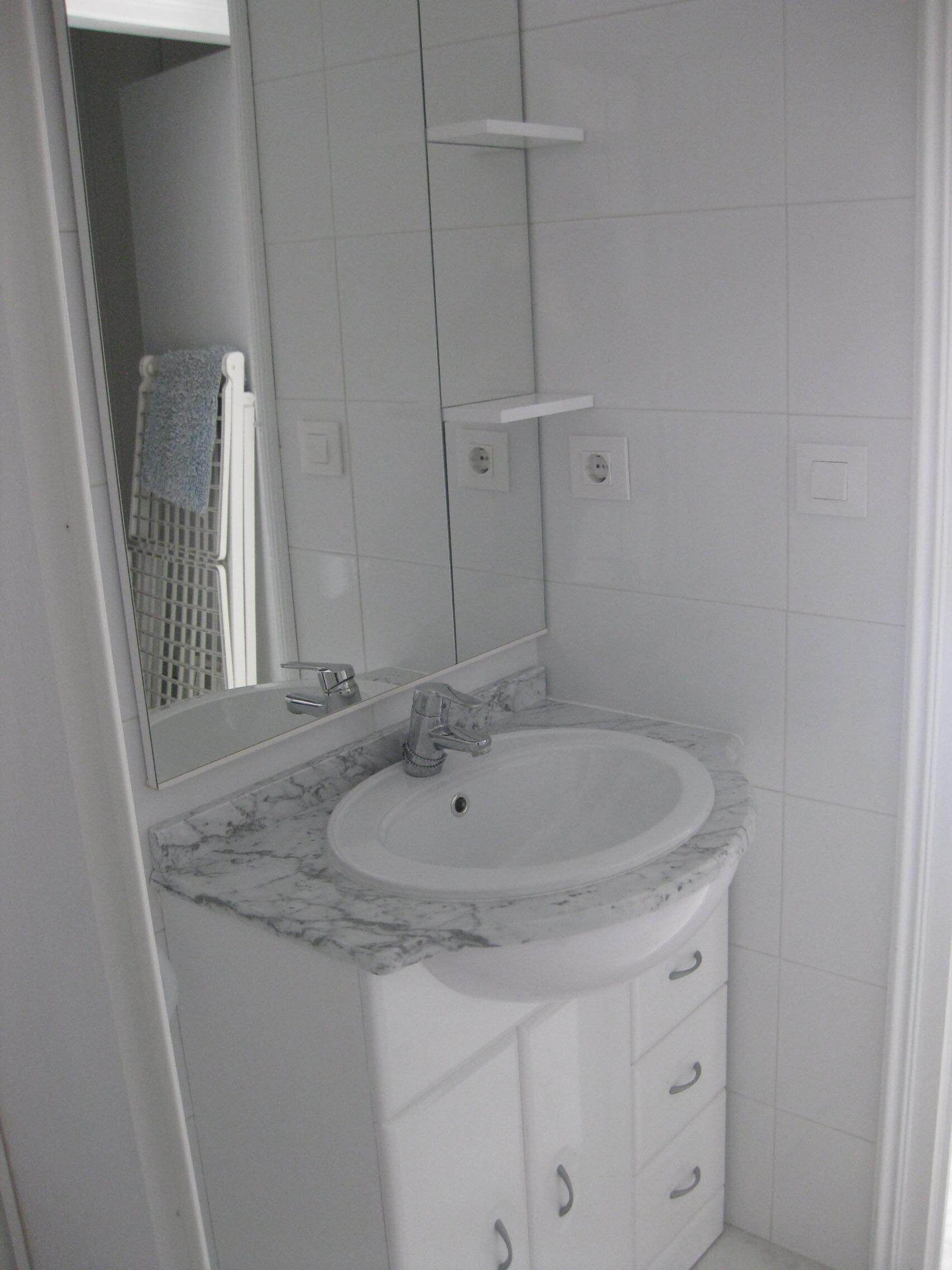 Une salle de bain avec un lavabo blanc, un comptoir en marbre et un miroir au-dessus. Un porte-serviettes est fixé au mur et, sous le lavabo, on trouve un meuble blanc.