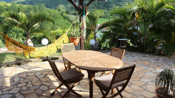 Een overdekt terras met een stenen vloer, een houten tafel met vier stoelen, een hangmat en potplanten.