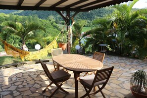 Une terrasse couverte avec un sol en pierre, une table en bois et quatre chaises, un hamac et des plantes en pot.