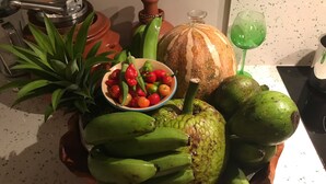 Een houten dienblad met bananen, een kom rode en groene paprika's, een grote groene vrucht en een groen glas.