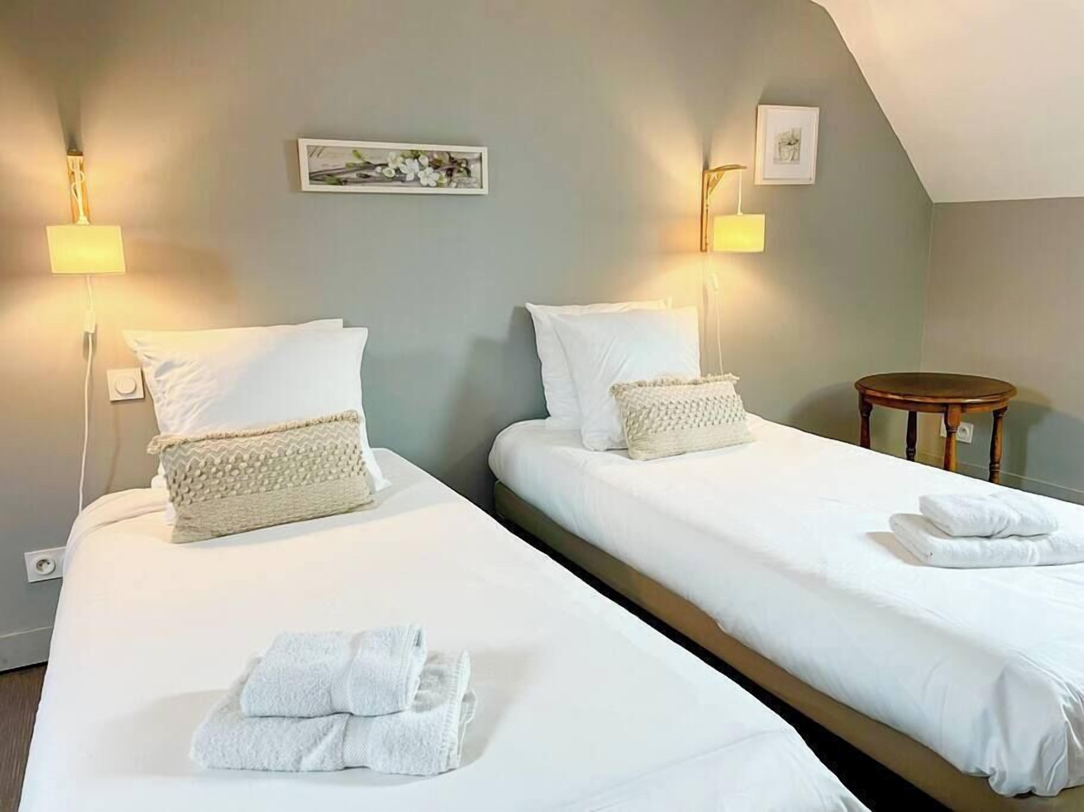 A hotel room with two beds, each with white linens and pillows, a small round wooden table, and wall-mounted lamps.