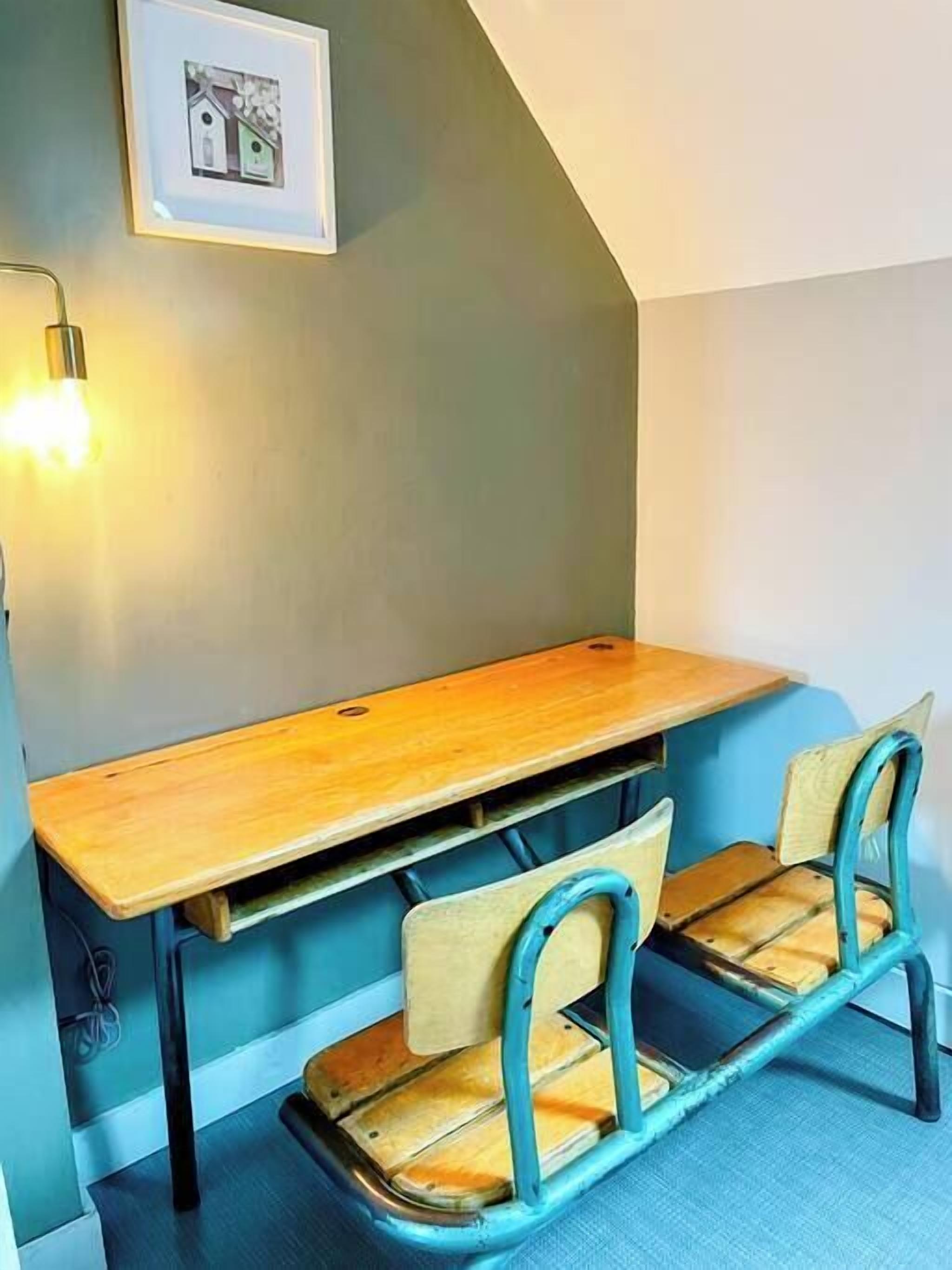 Una habitación pequeña y acogedora con un escritorio de madera y dos sillas, un cuadro en la pared y una lámpara.