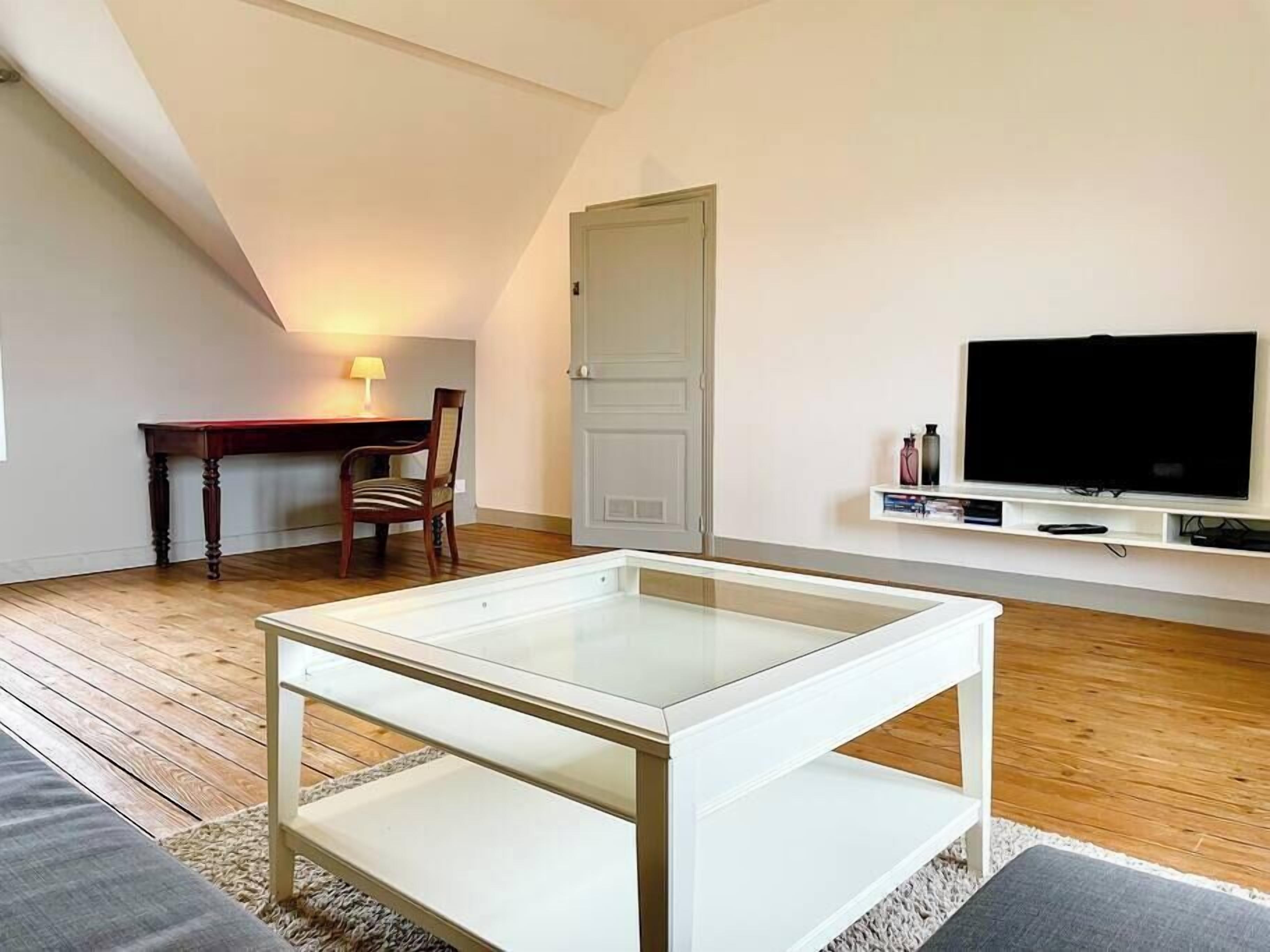 A modern living room with a glass-top coffee table, a wooden desk with a chair, a television on a white shelf, and a door in the background.