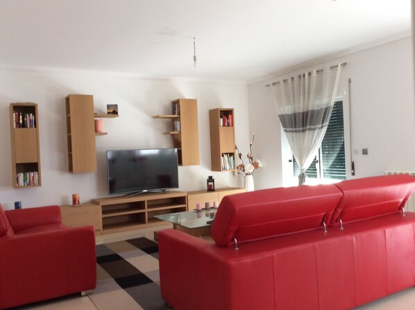 A living room with a flat-screen TV, red sofas, wooden bookshelves, and a glass coffee table.