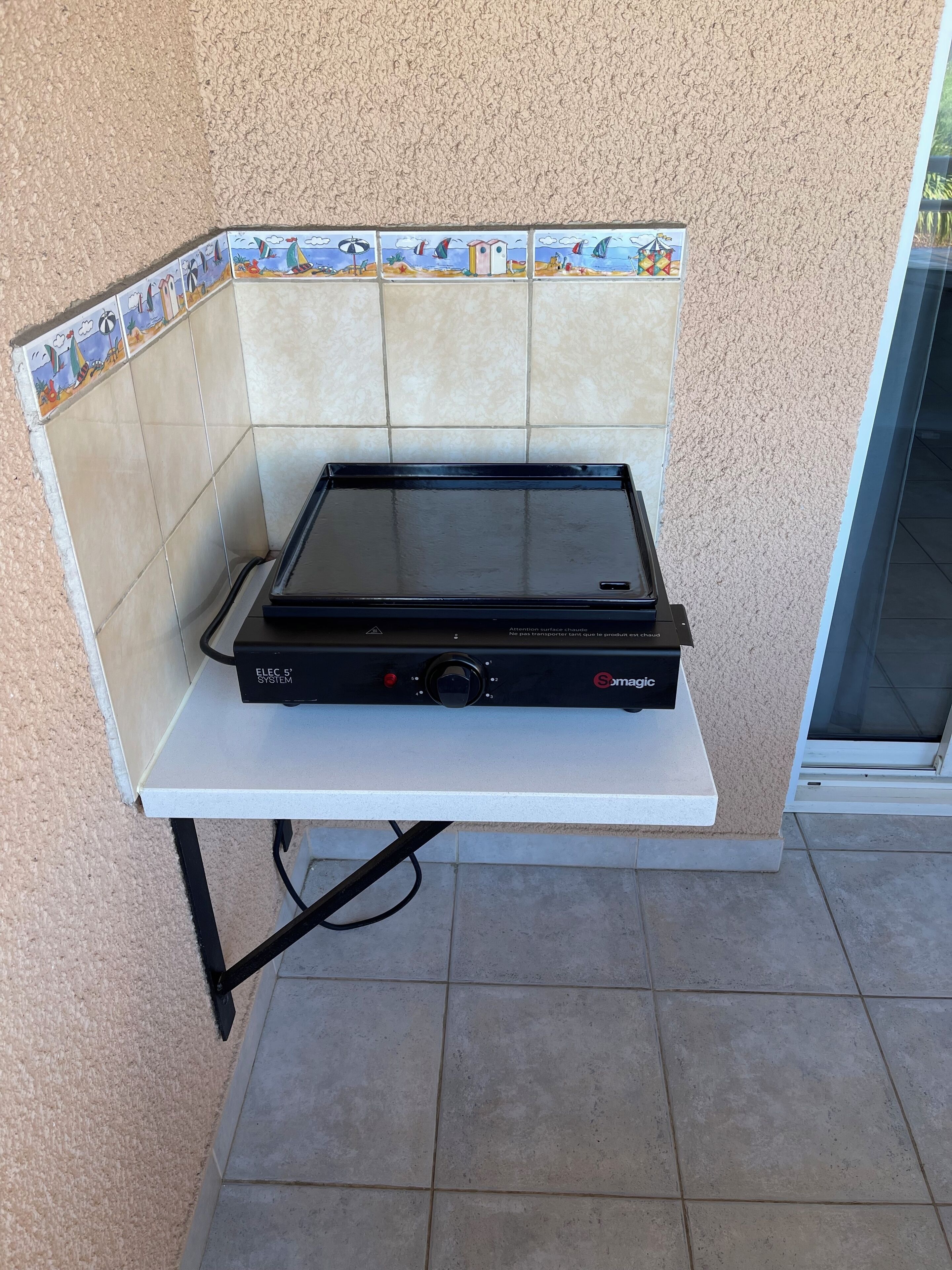 A portable electric grill on a white table against a textured wall with a tiled backsplash.