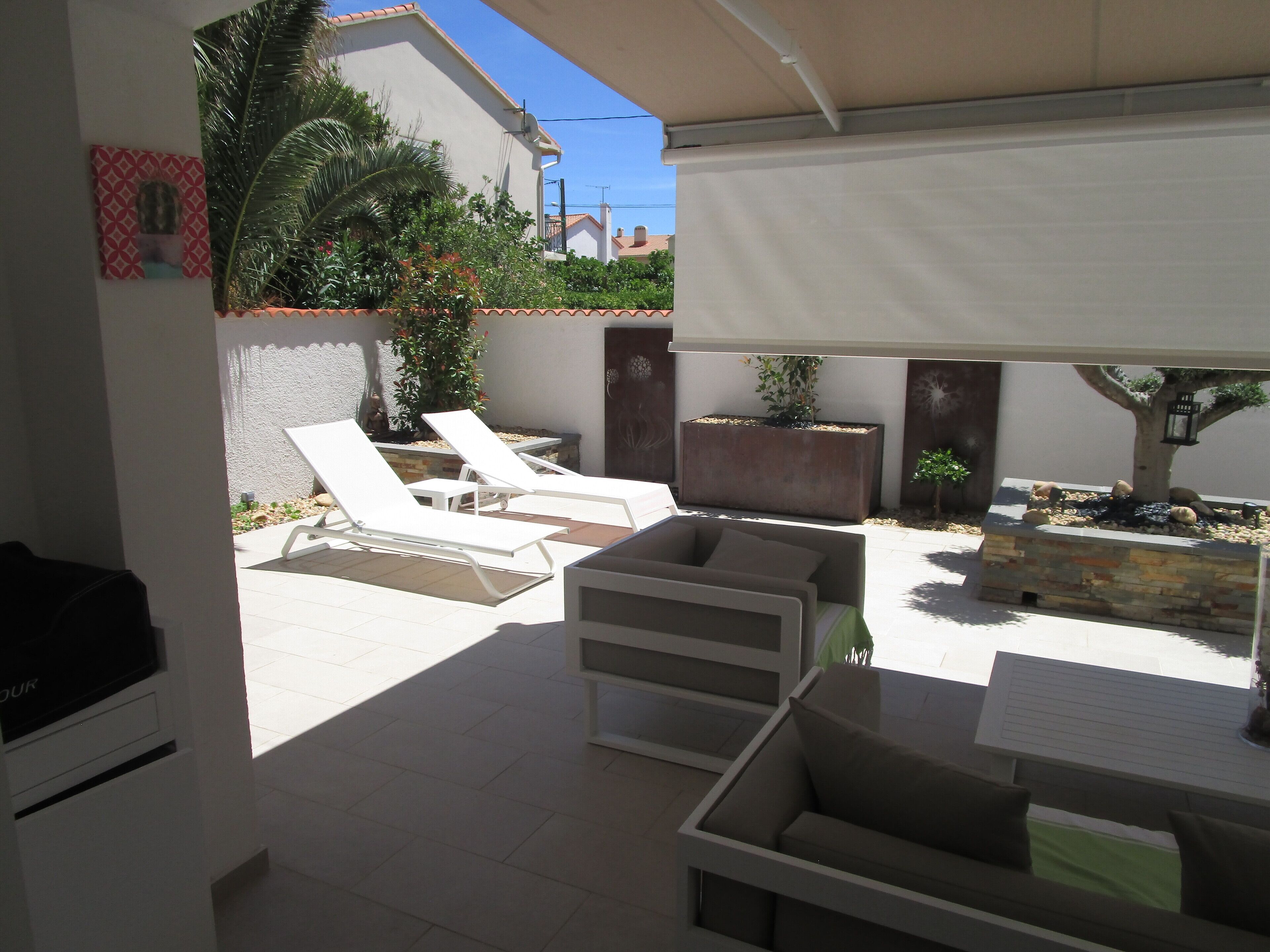 A sunlit patio with white lounge chairs, a stone fireplace, and a small tree.
