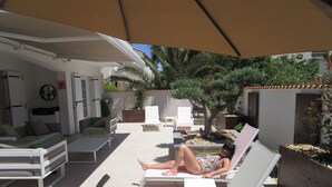 Un espace de détente extérieur baigné de soleil, avec des chaises longues blanches, une table et un grand parasol pour créer de l’ombre.