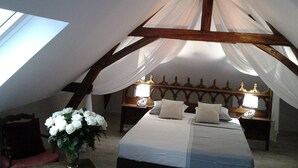 Une chambre avec un plafond haut, un lit avec une literie blanche, deux lampes de chevet et un vase de fleurs blanches posé sur une table de chevet.