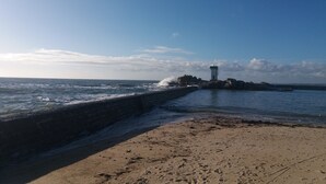 Un paysage côtier avec un phare, des vagues qui s’écrasent contre un mur de pierre et une plage de sable.