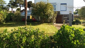 Une maison à deux étages, à l’extérieur blanc et aux bordures bleues, entourée d’un jardin bien entretenu où poussent diverses plantes et un palmier.