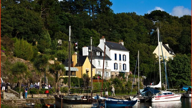 Un port où sont amarrés des bateaux, une maison blanche aux volets bleus et un escalier en pierre qui descend vers l’eau.