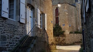 Una estrecha calle adoquinada bordeada por edificios de piedra con contraventanas blancas, una escalera de metal y un camino empedrado que conduce a un edificio con un ventanal rojo.