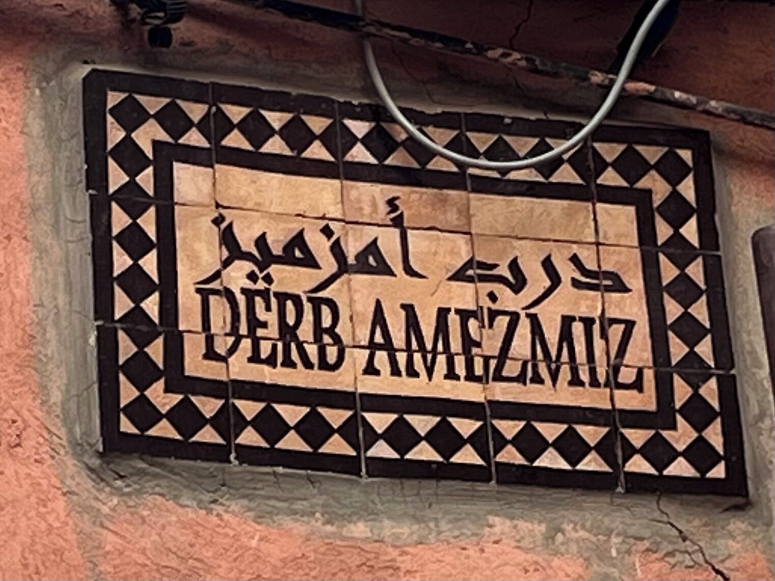 Une enseigne portant le nom « DERB AMEZMIZ » en arabe et en anglais, entourée d’un motif géométrique de carreaux.