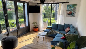 Ein modernes Wohnzimmer mit einem blauen Sofa, einem schwarzen Pouf und einem Couchtisch. Ein großes Fenster bietet Blick auf Grünflächen, und an der Wand hängt ein Gemälde.