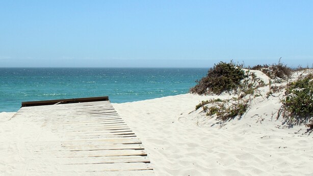 Eine hölzerne Promenade führt zu einem Sandstrand mit klarem, blauem Wasser und wolkenlosem Himmel.