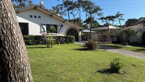 Una casa con un césped cuidado, una pérgola con sillas y un gran árbol en primer plano.