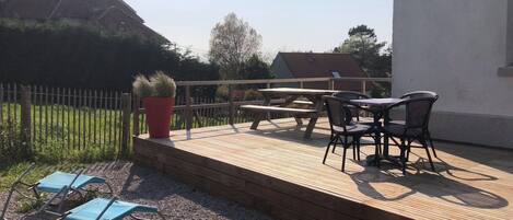 Une terrasse en bois avec une table et des chaises, un plant en pot rouge et une chaise pliante bleue.