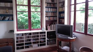 Um quarto com uma janela grande, uma estante cheia de livros, uma televisão sobre um suporte e um armário de madeira.