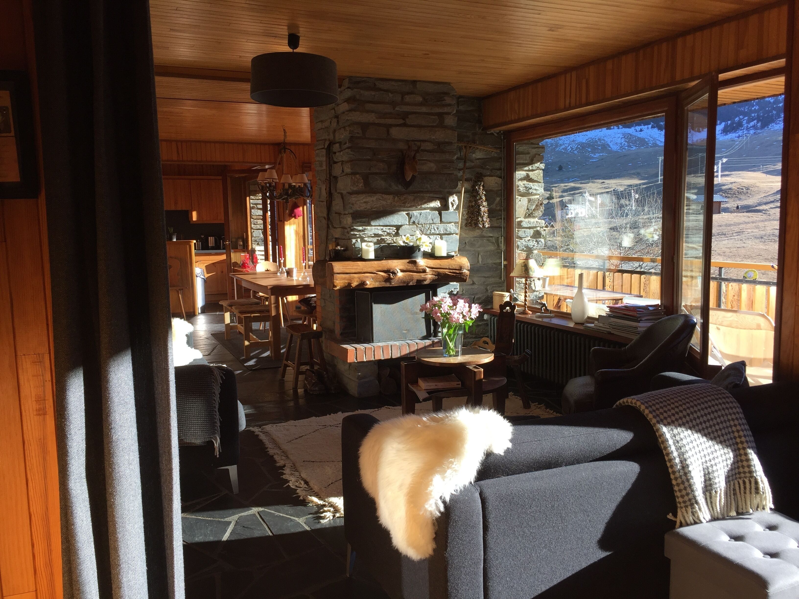 A cozy living room with a stone fireplace, wooden furniture, and a view of a mountainous landscape through large windows.