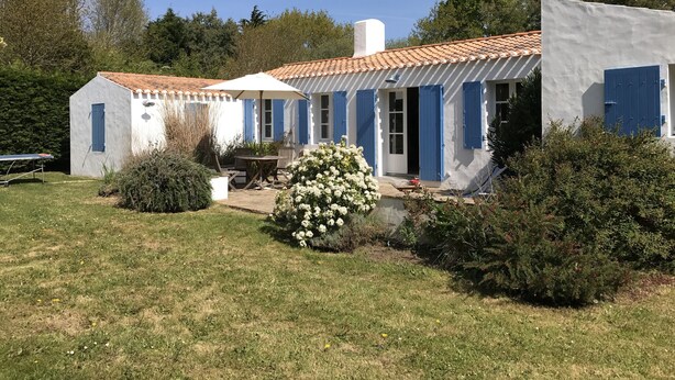 Ein weißes Gebäude mit blauen Fensterläden, eine Terrasse mit Tisch und Stühlen sowie ein kleiner Garten mit weißen Blüten.