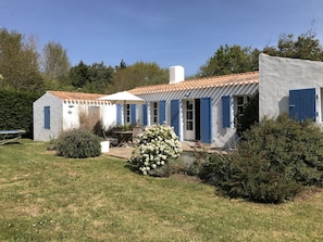 Un edificio blanco con contraventanas azules, un patio con mesa y sillas, y un pequeño jardín con flores blancas.