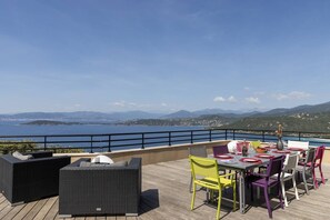Terraço com área externa para sentar e mesa posta para refeições, com vista para um corpo d'água e montanhas ao longe.