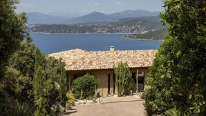 A villa with a tiled roof, surrounded by cacti and trees, overlooking a lake and mountains.