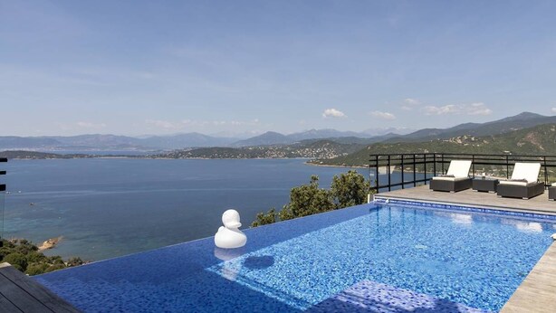 Une piscine à débordement offrant une vue imprenable sur la mer et les montagnes, des chaises longues et un objet décoratif blanc.