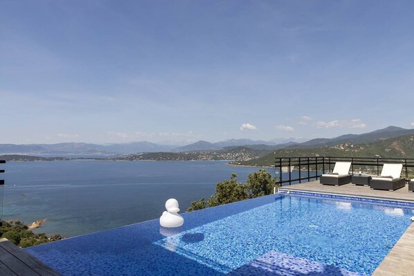 An infinity pool with a clear view of the sea and mountains, lounge chairs, and a white decorative object.
