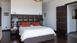 Une chambre à coucher avec une tête de lit capitonnée, un lit recouvert de linge de lit blanc et agrémenté d’oreillers orange, des lampes de chevet, un plancher en bois et une porte avec un panneau foncé.