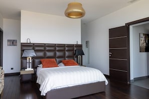A bedroom with a tufted headboard, a bed with white bedding and orange pillows, bedside lamps, a wooden floor, and a door with a dark panel.