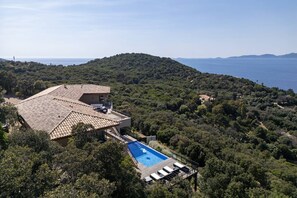 A villa with a swimming pool, surrounded by dense forest and overlooking a coastal area.