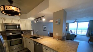 A modern kitchen with stainless steel appliances, a granite countertop, and a view of the ocean through the living area. - Shore to Please beach lovers paradise! (New Smyrna Beach)