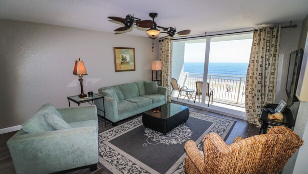 A living room with a sofa, armchair, and a coffee table. There is a ceiling fan, a lamp, and a view of the ocean through large windows. - Shore to Please beach lovers paradise! (New Smyrna Beach)