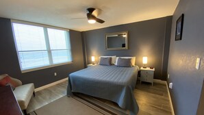 A bedroom with a bed, bedside tables, a ceiling fan, a window with blinds, a mirror, and a rug. - Shore to Please beach lovers paradise! (New Smyrna Beach)