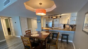 A dining area with a wooden table and chairs, a pendant light, and a kitchen in the background. - Shore to Please beach lovers paradise! (New Smyrna Beach)