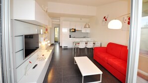 Un salon moderne avec un canapé rouge, une table basse blanche et un téléviseur posé sur un meuble blanc. On y trouve également un coin cuisine avec des meubles blancs et une table à manger avec des chaises.