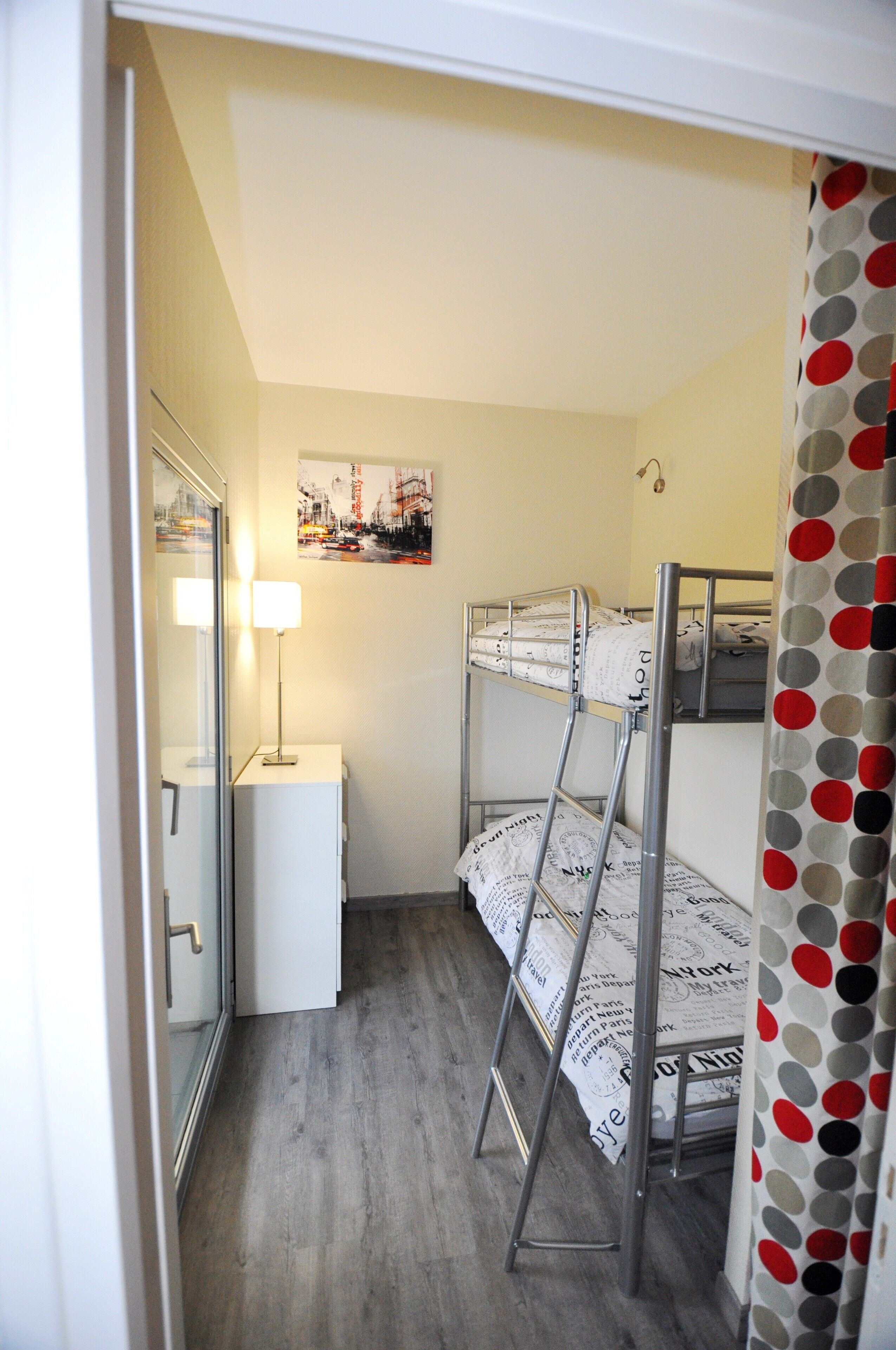 A small, narrow room with bunk beds, a desk, a lamp, and a mirror.