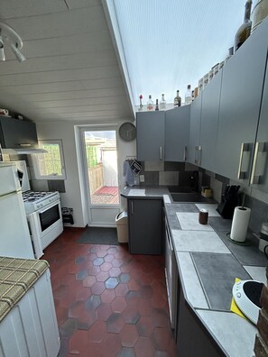 A compact kitchen with a gas stove, oven, and a small refrigerator. There is a sink, a countertop with a knife block, and a window providing natural light.