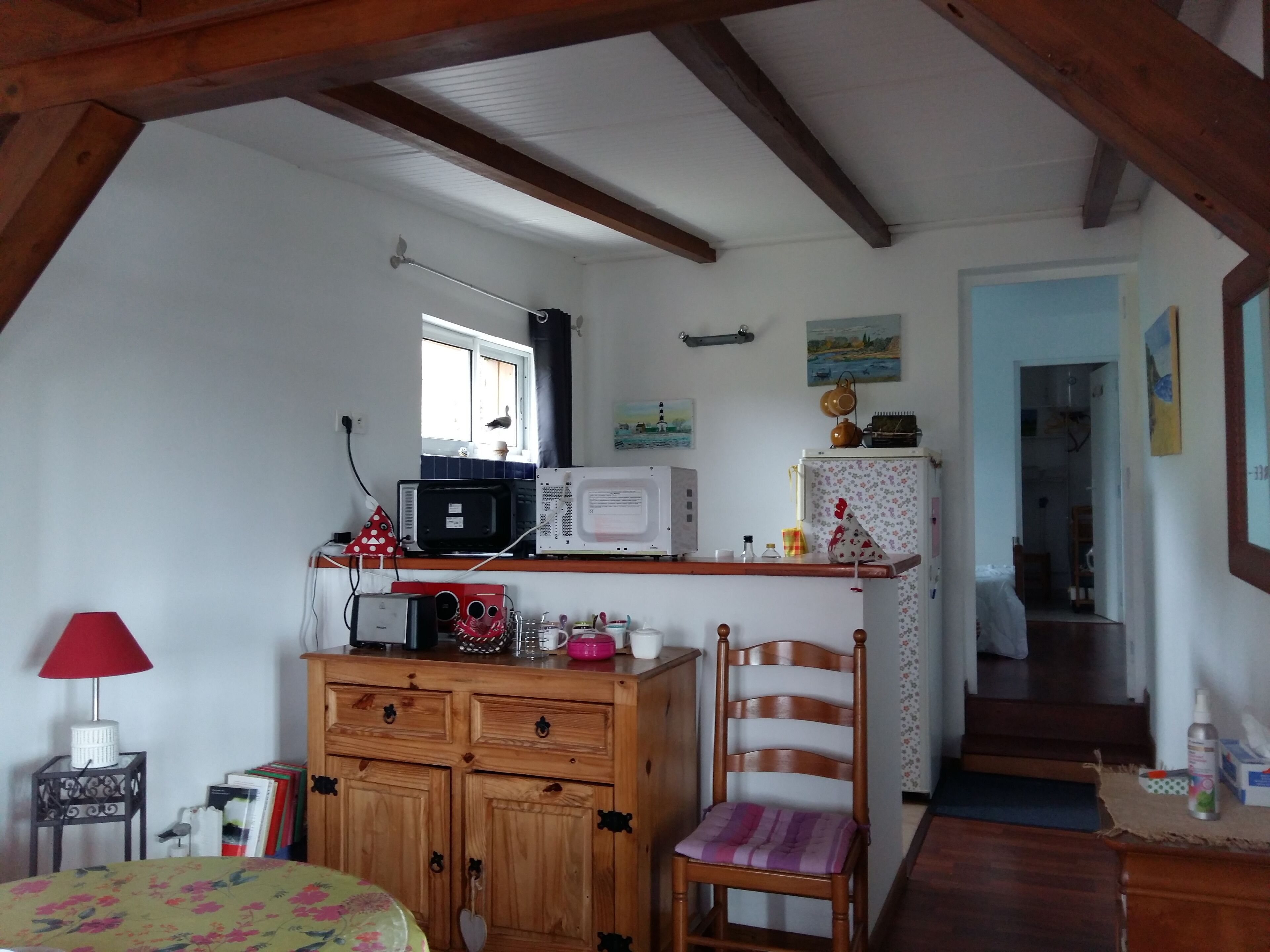 Une cuisine chaleureuse avec des meubles en bois, un four à micro-ondes et un réfrigérateur. Une fenêtre laisse entrer la lumière naturelle, et il y a une chaise avec un coussin.